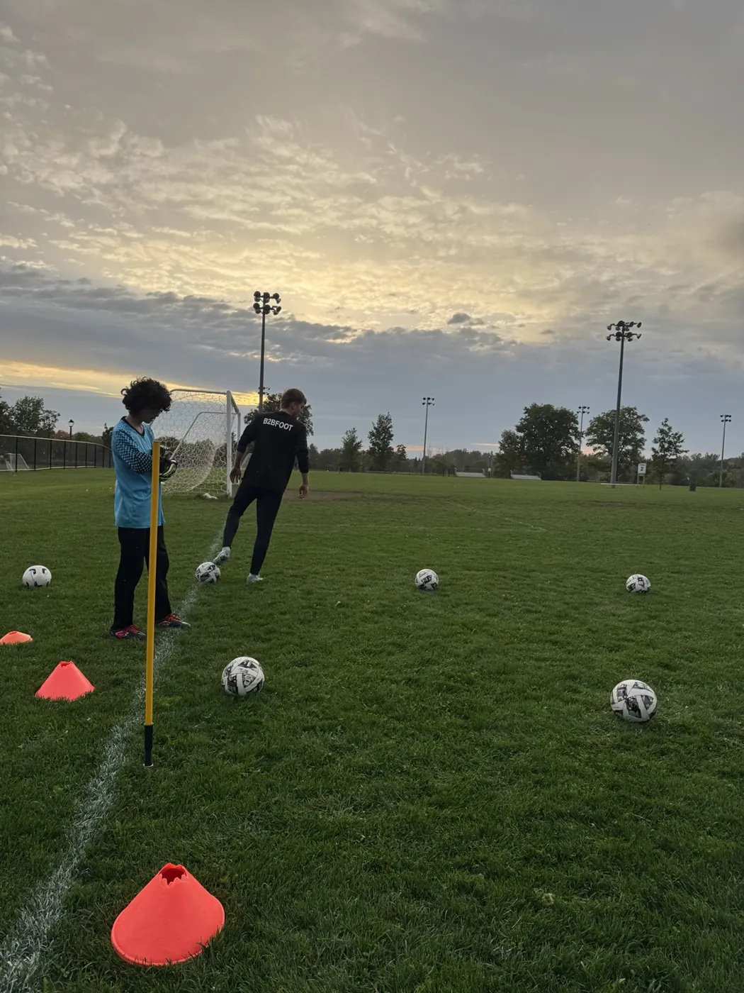 B2B coach and goalkeeper practicing with balls and cones on the field at sunset